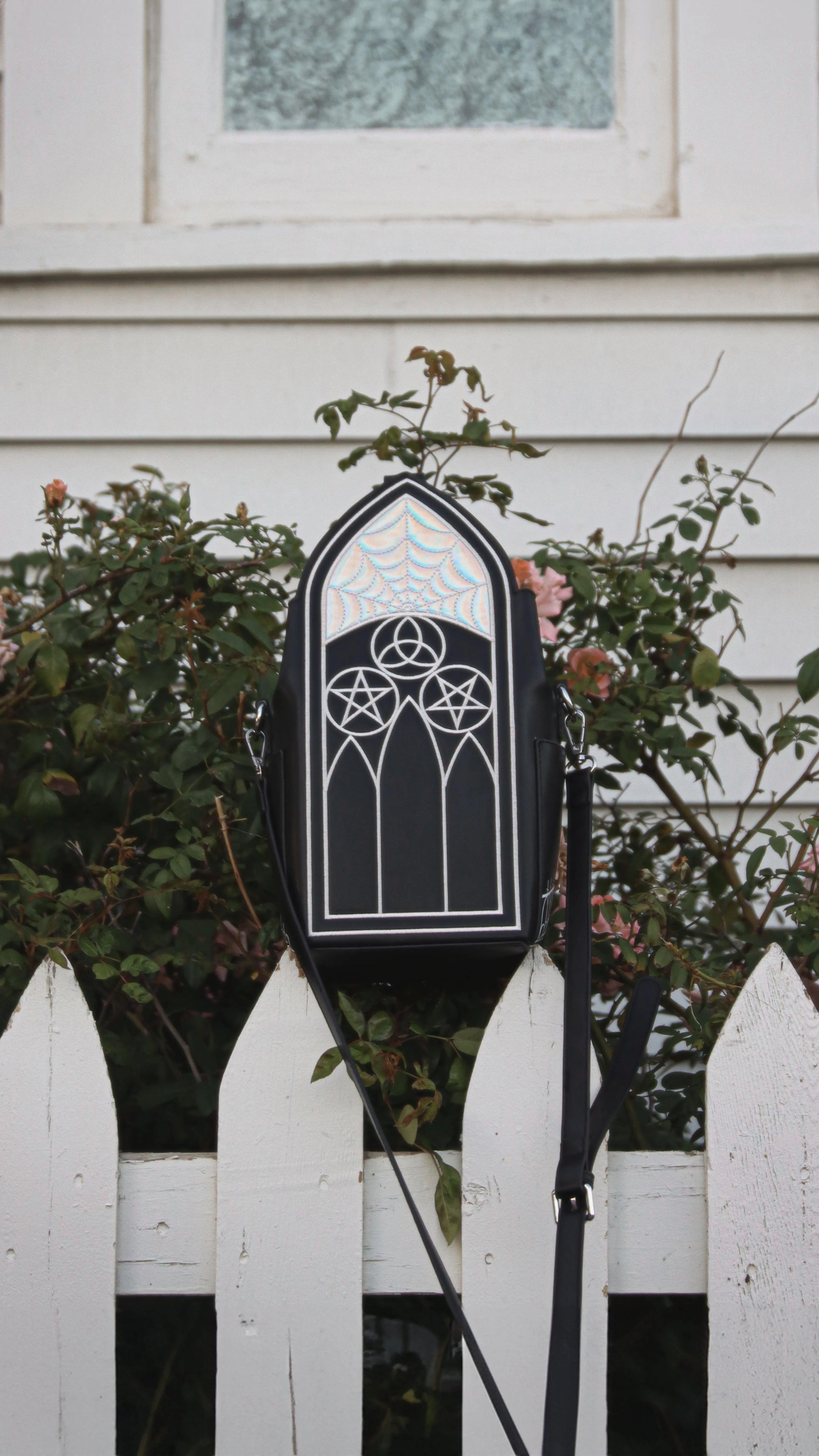 A black purse featuring Gothic inspired architecture. The top has a holographic spiderweb design. The bag is sitting on top of a white fence outside.