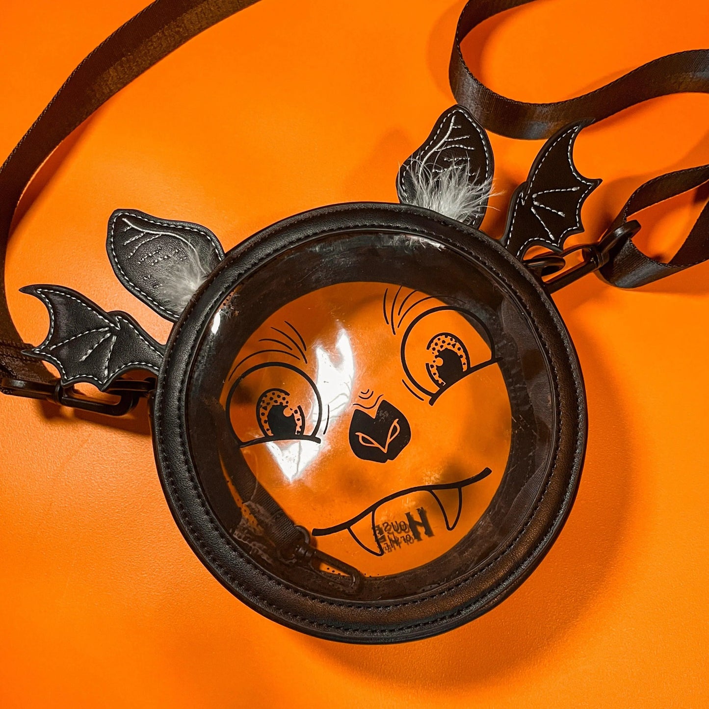 A Halloween purse featuring a whimsical bat face design with a clear center, long black strap, and ears that have white feathers. The bag is sitting on an orange background.
