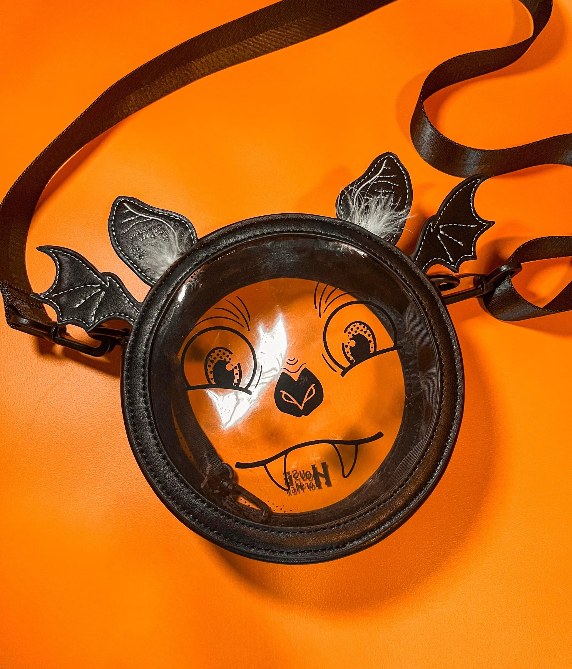 A Halloween purse featuring a whimsical bat face design with a clear center, long black strap, and ears that have white feathers. The bag is sitting on an orange background.