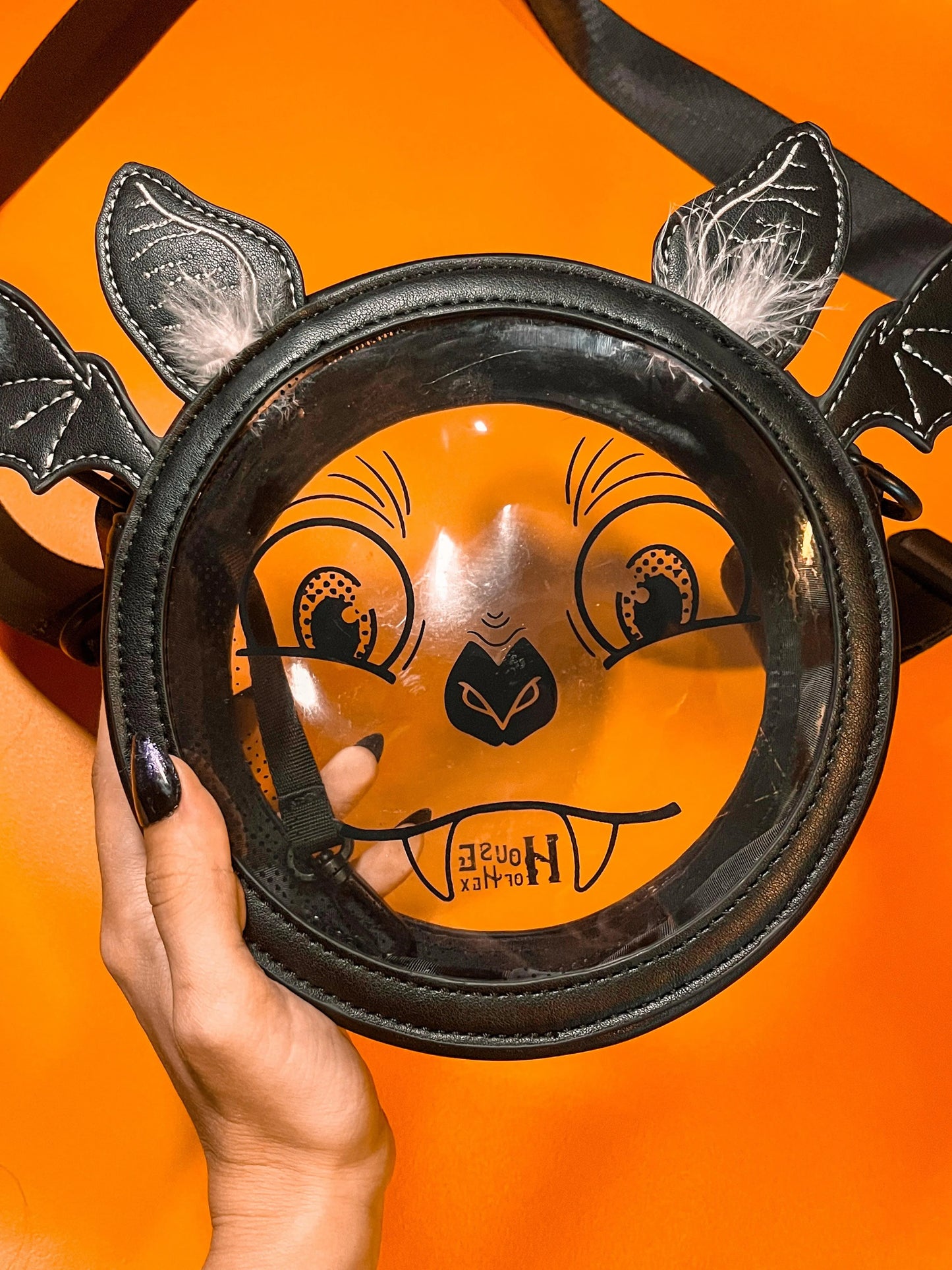A Halloween purse featuring a whimsical bat face design with a clear center, long black strap, and ears that have white feathers. The bag is sitting on an orange background.
