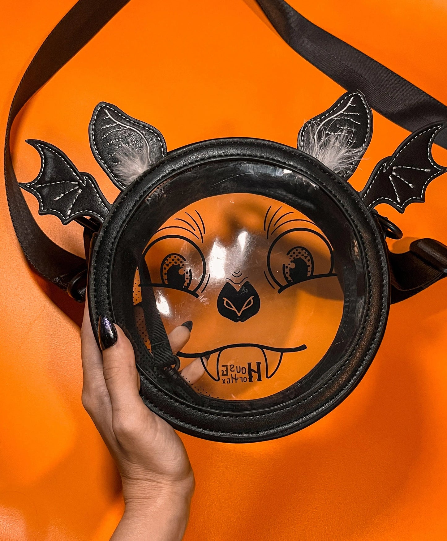 A Halloween purse featuring a whimsical bat face design with a clear center, long black strap, and ears that have white feathers. The bag is sitting on an orange background.
