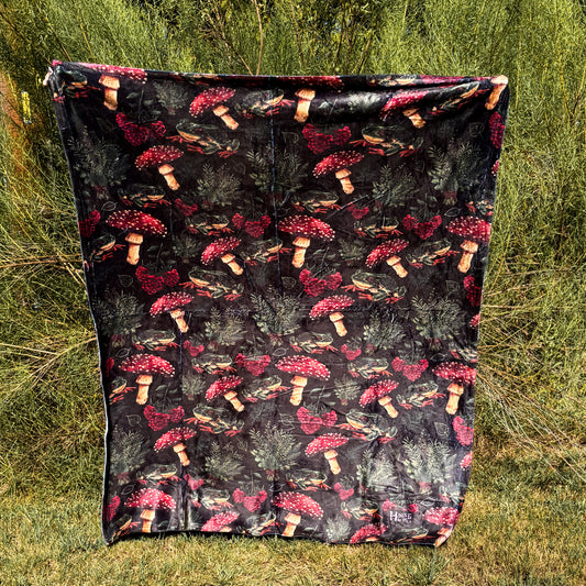 A decorative blanket featuring a pattern of mushrooms, plants, and frogs on a black background.