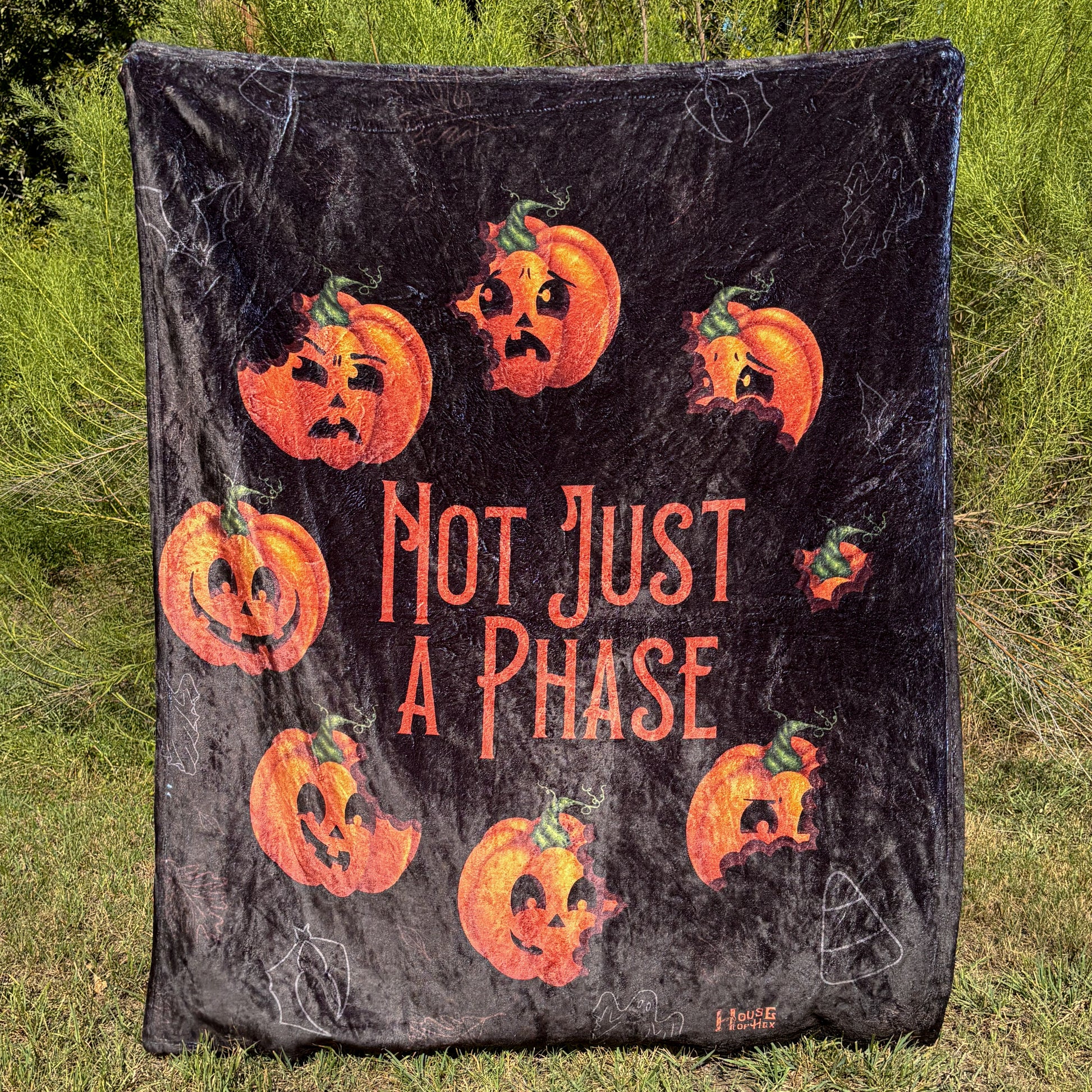 A black blanket featuring designs pumpkins in a circle with different facial expressions, in the middle in orange font it says "Not Just A Phase".