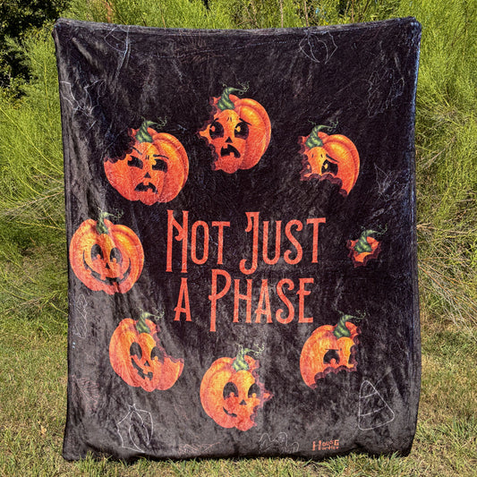A black blanket featuring designs pumpkins in a circle with different facial expressions, in the middle in orange font it says "Not Just A Phase".