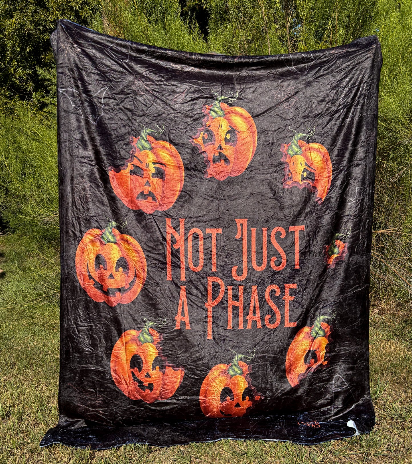 A black blanket featuring designs pumpkins in a circle with different facial expressions, in the middle in orange font it says "Not Just A Phase".