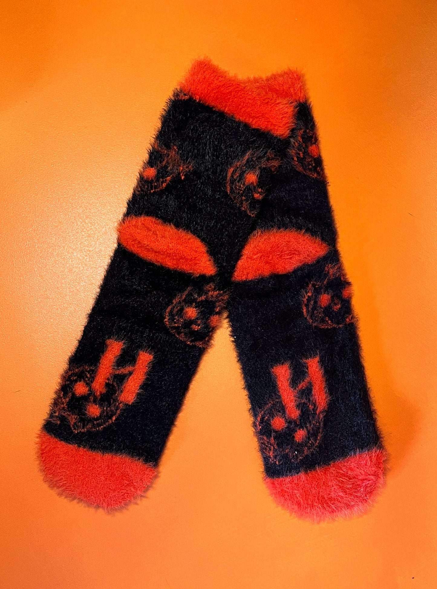 Black fuzzy socks with orange on the toe, heel, and ankle featuring a pattern of pumpkin shaped cat heads.
