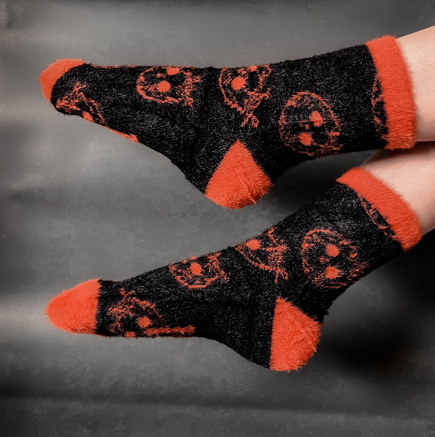 Black fuzzy socks with orange on the toe, heel, and ankle featuring a pattern of pumpkin shaped cat heads.