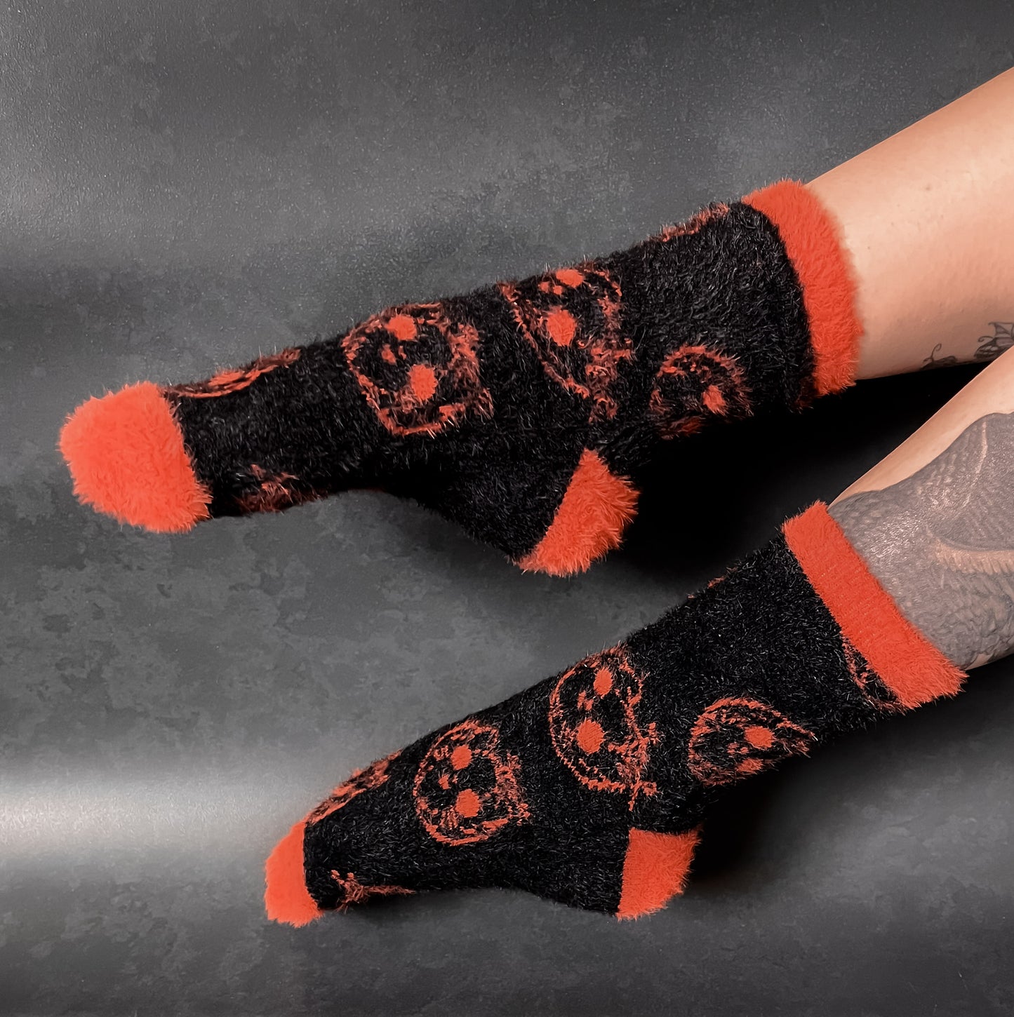 Black fuzzy socks with orange on the toe, heel, and ankle featuring a pattern of pumpkin shaped cat heads.