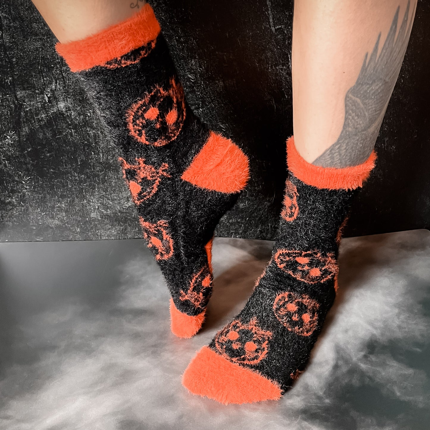 Black fuzzy socks with orange on the toe, heel, and ankle featuring a pattern of pumpkin shaped cat heads.