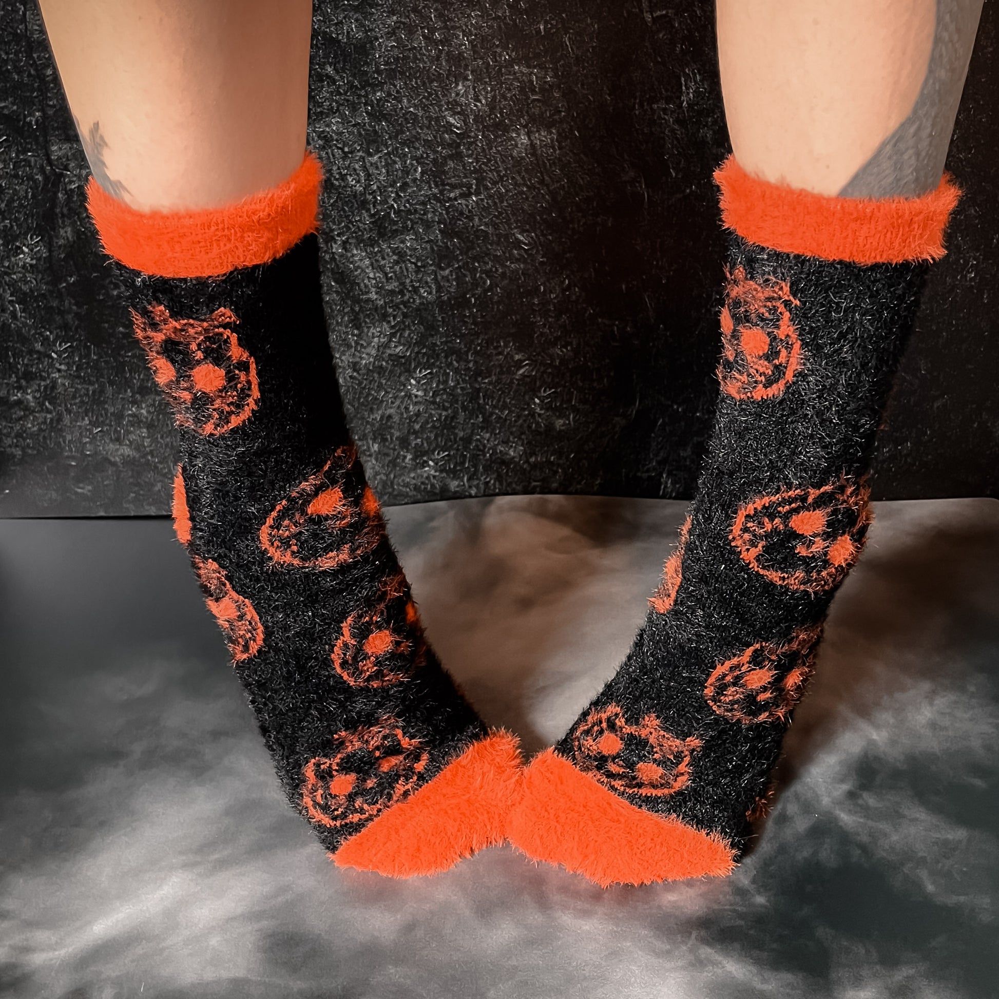 Black fuzzy socks with orange on the toe, heel, and ankle featuring a pattern of pumpkin shaped cat heads.