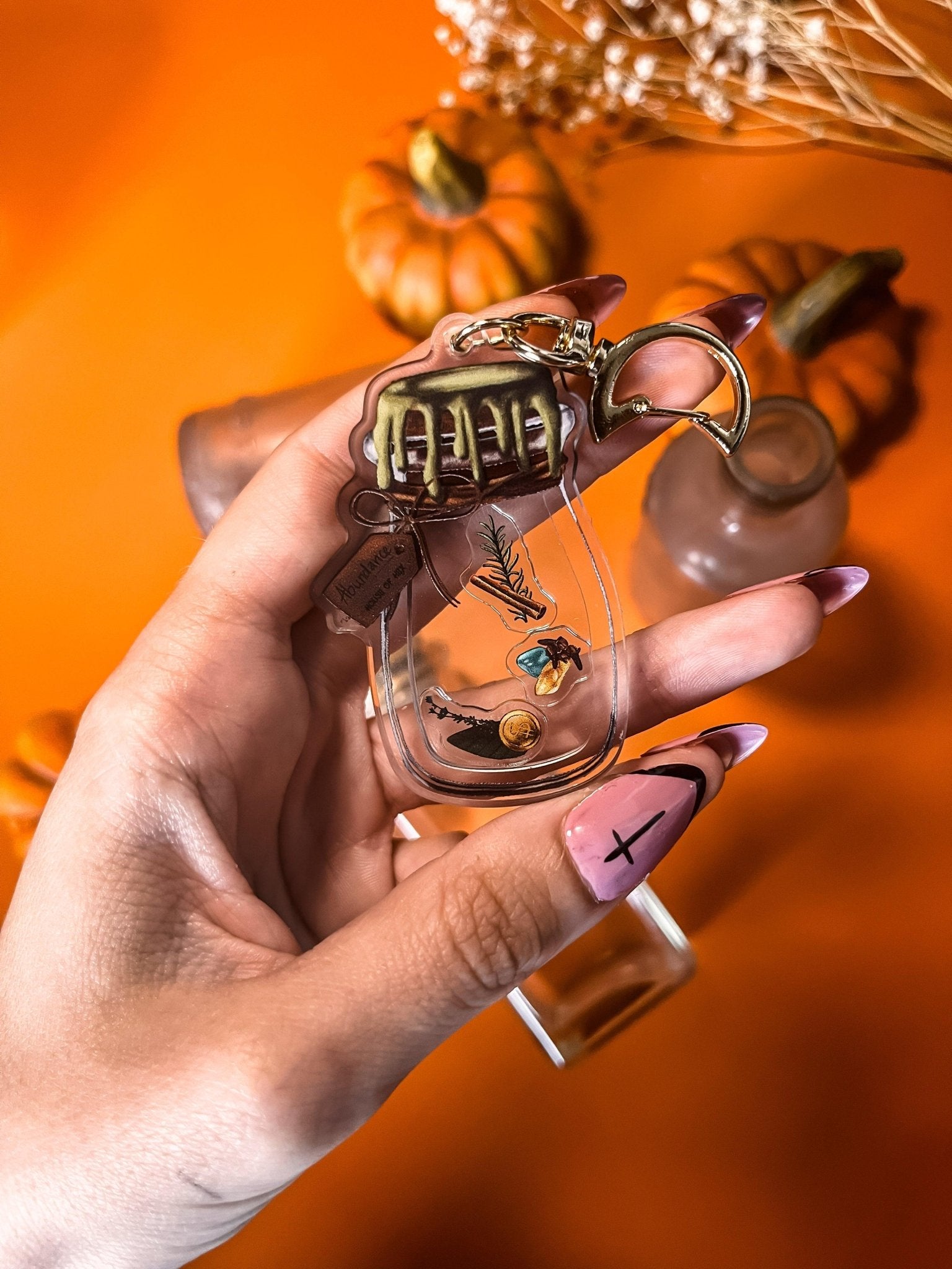 A decorative keychain displaying a small jar filled with cinnamon, bay leaves, coins, cloves, citrine, aventurine, and rosemary with a gold top.