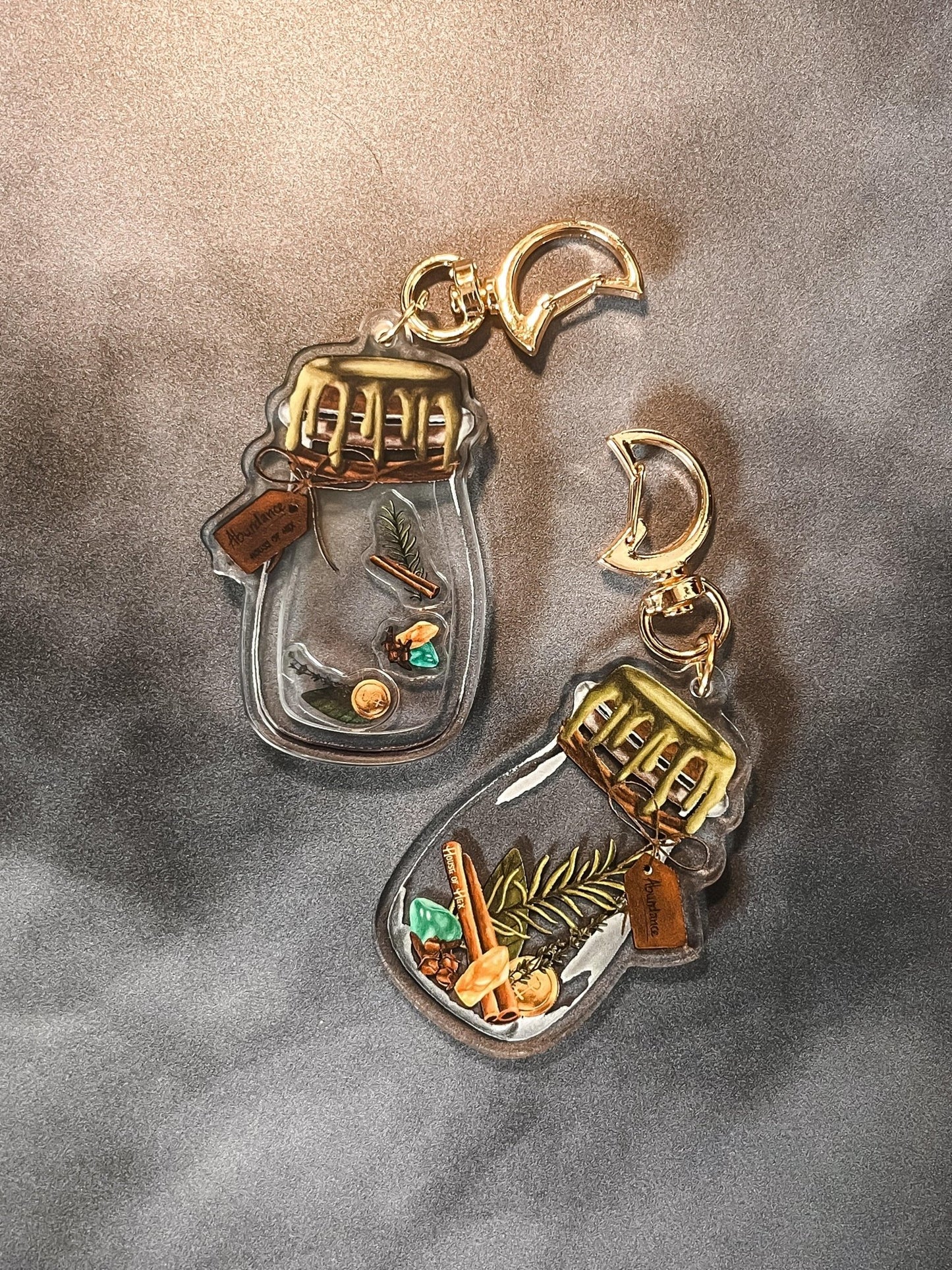 Two decorative keychains displaying a small jar filled with cinnamon, bay leaves, coins, cloves, citrine, aventurine, and rosemary with a gold top.