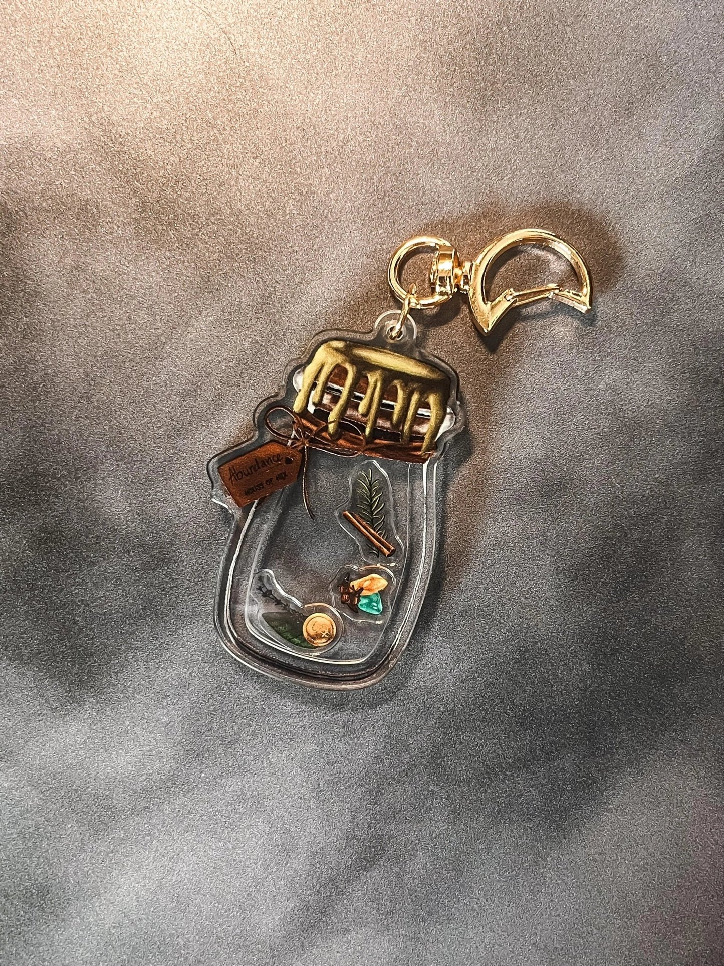 A decorative keychain displaying a small jar filled with cinnamon, bay leaves, coins, cloves, citrine, aventurine, and rosemary with a gold top.