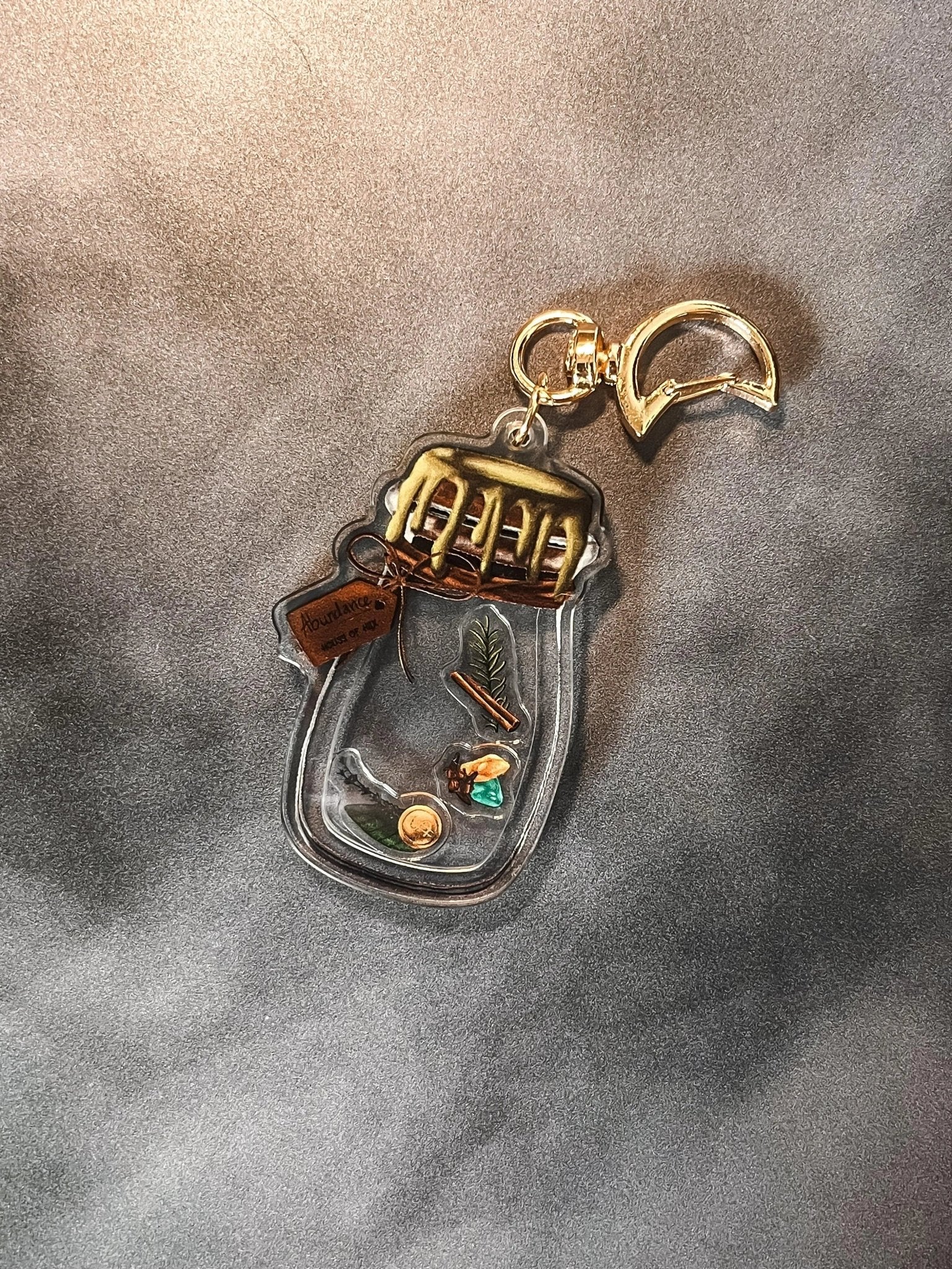 A decorative keychain displaying a small jar filled with cinnamon, bay leaves, coins, cloves, citrine, aventurine, and rosemary with a gold top.