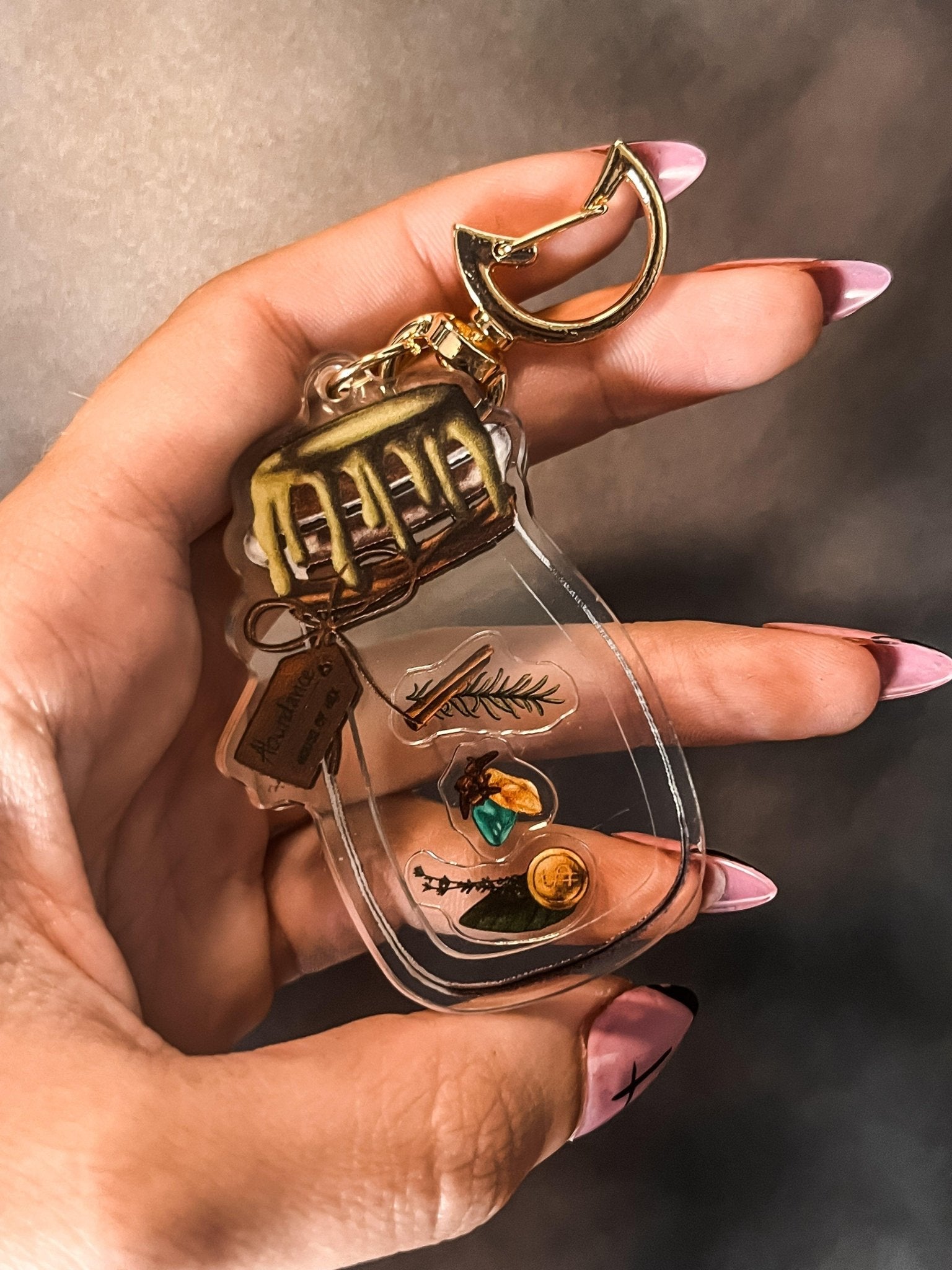 A decorative keychain displaying a small jar filled with cinnamon, bay leaves, coins, cloves, citrine, aventurine, and rosemary with a gold top.