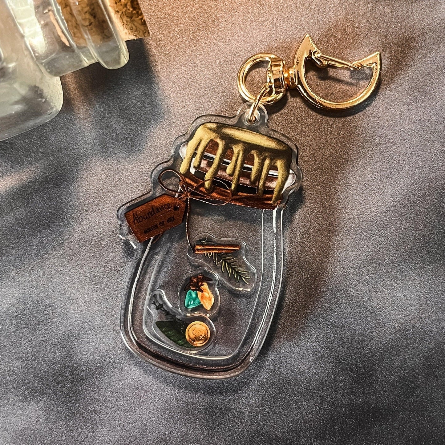 A decorative keychain displaying a small jar filled with cinnamon, bay leaves, coins, cloves, citrine, aventurine, and rosemary with a gold top.
