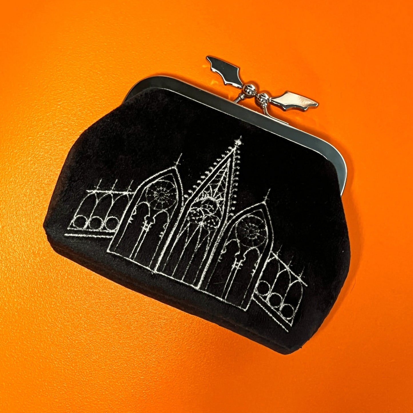 Black coin purse featuring gothic gables. The clasp of the purse in the shape of a bat. The bag is on top of an orange background.