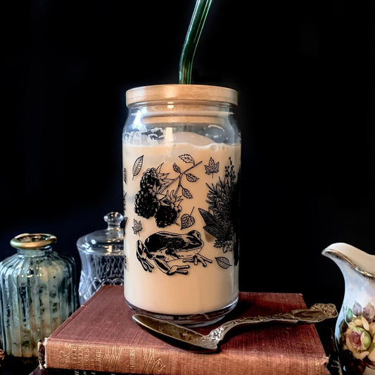 A glass drinking jar with a bamboo lid and green glass straw, the glass features designs of mushrooms, frogs, and leaves.