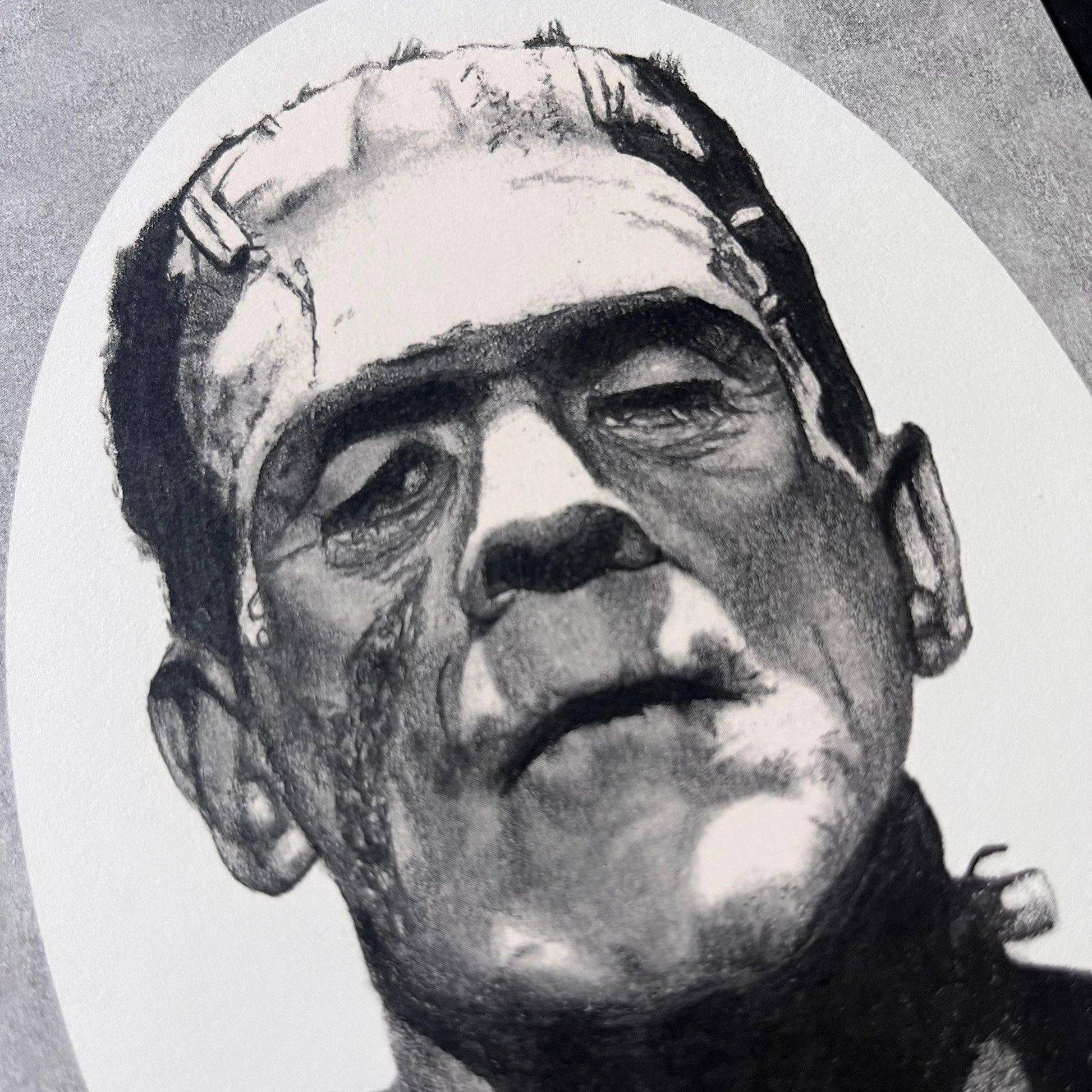 Black and white portrait drawing of Frankenstein's monster on a white oval surrounded by a grey rectangle.