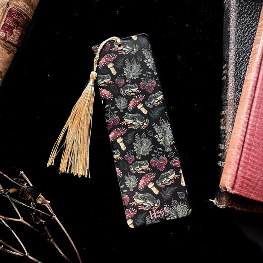A decorative bookmark featuring a pattern of mushrooms, plants, and frogs on a black background with a gold tassel.