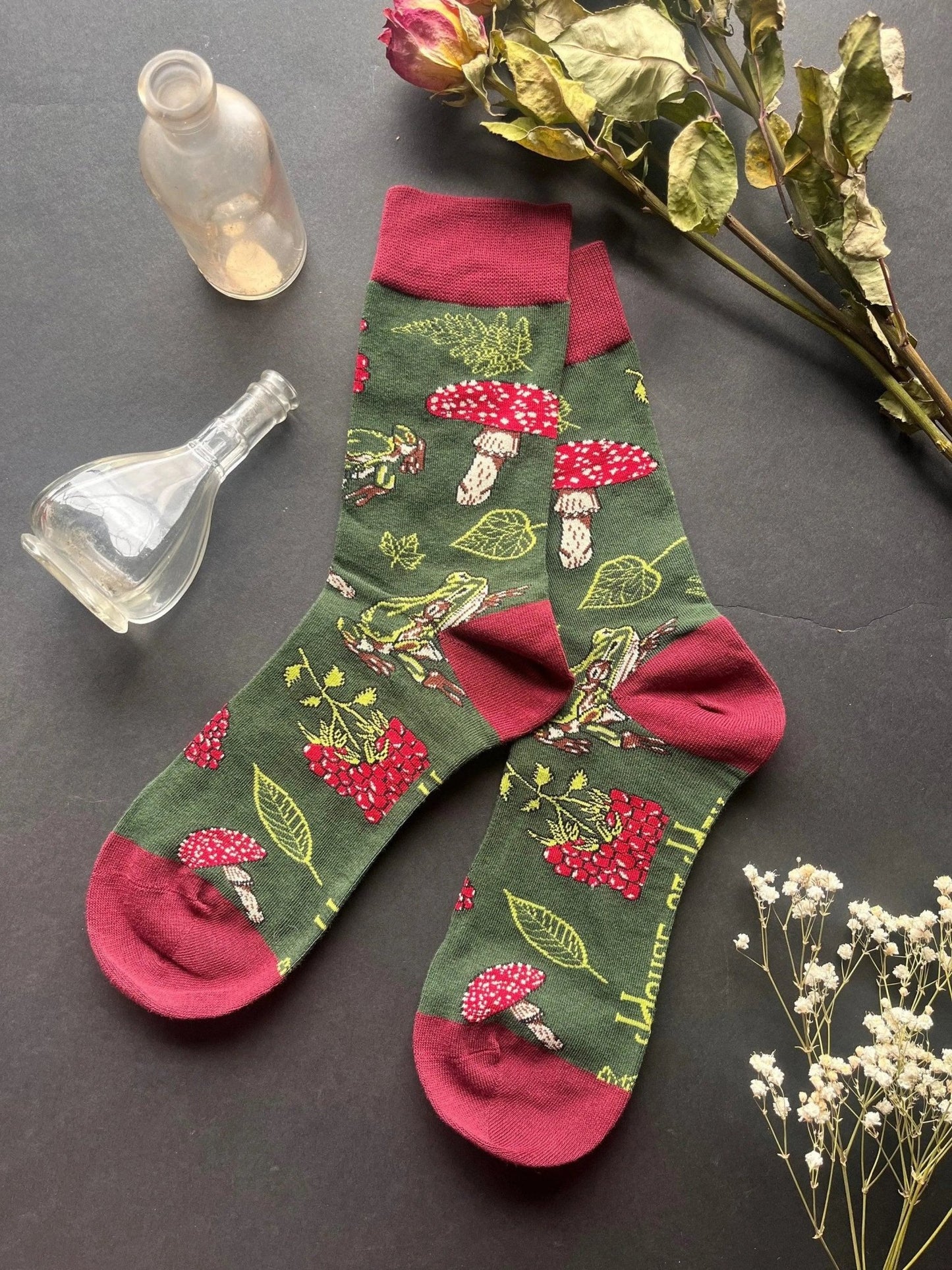 Socks with red on the toes, heel, and ankle section featuring a pattern of mushrooms, plants, and frogs on a green background.