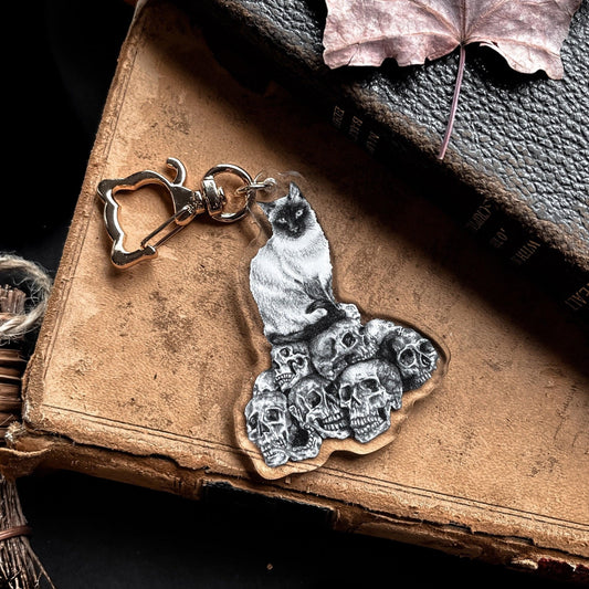 A black and white keychain with a drawing of a cat perched atop a collection of skulls with a golden cat-shaped clasp.