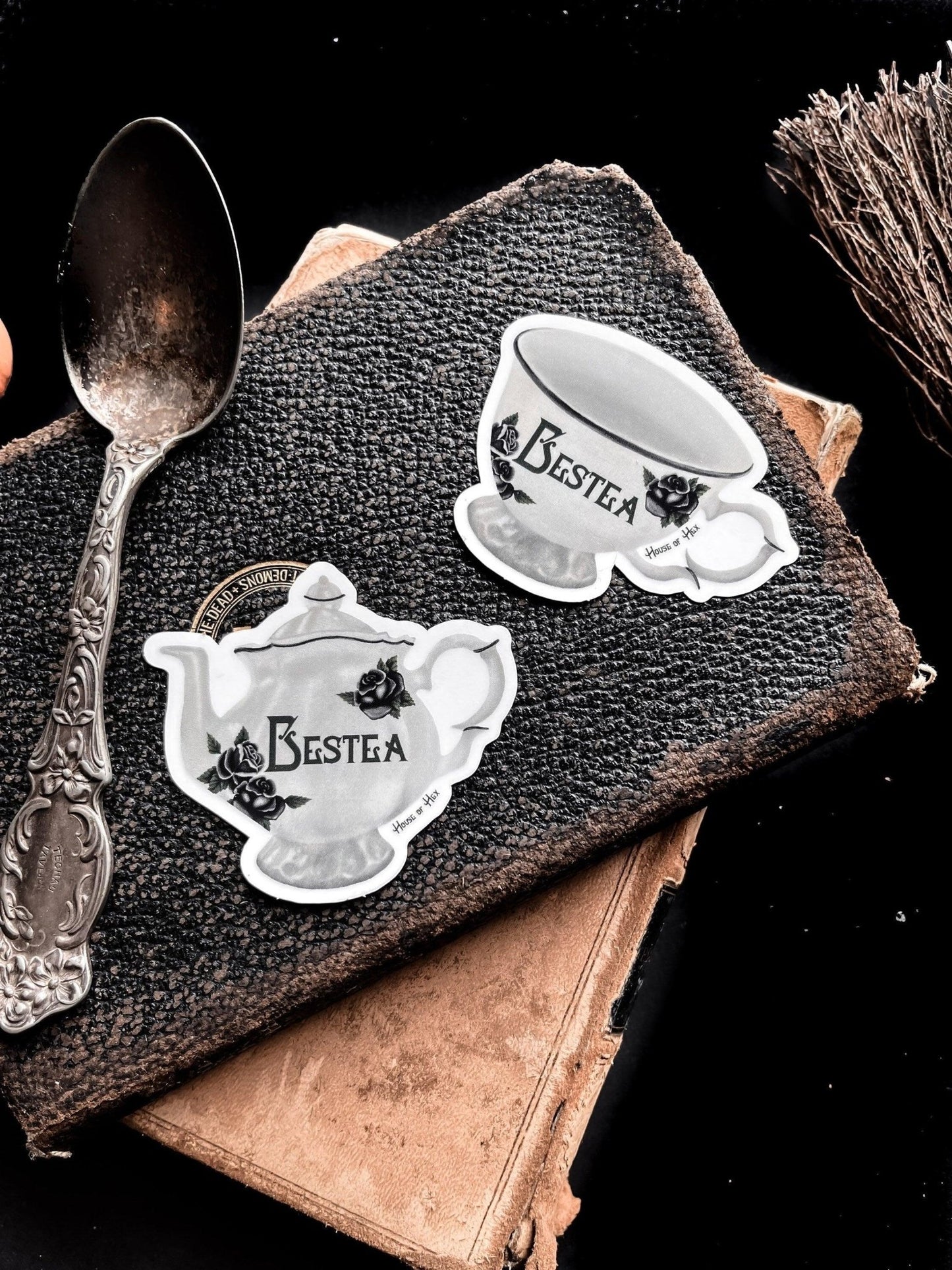 Two elegant white tea cup stickers with the word "Bestea" written in a gothic font placed on top of a leather notebook next to an antique silver spoon.