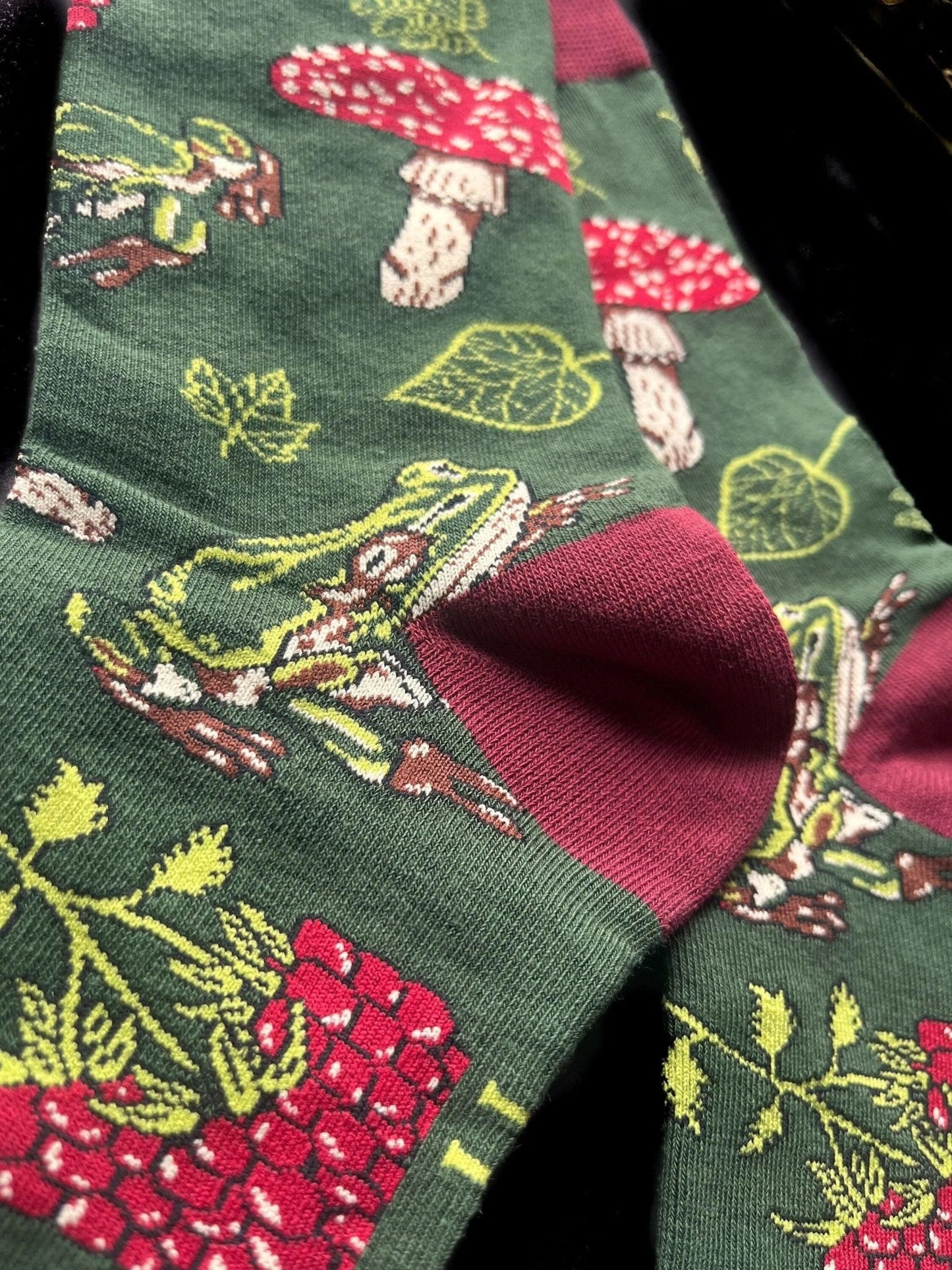 Socks with red on the toes, heel, and ankle section featuring a pattern of mushrooms, plants, and frogs on a green background.