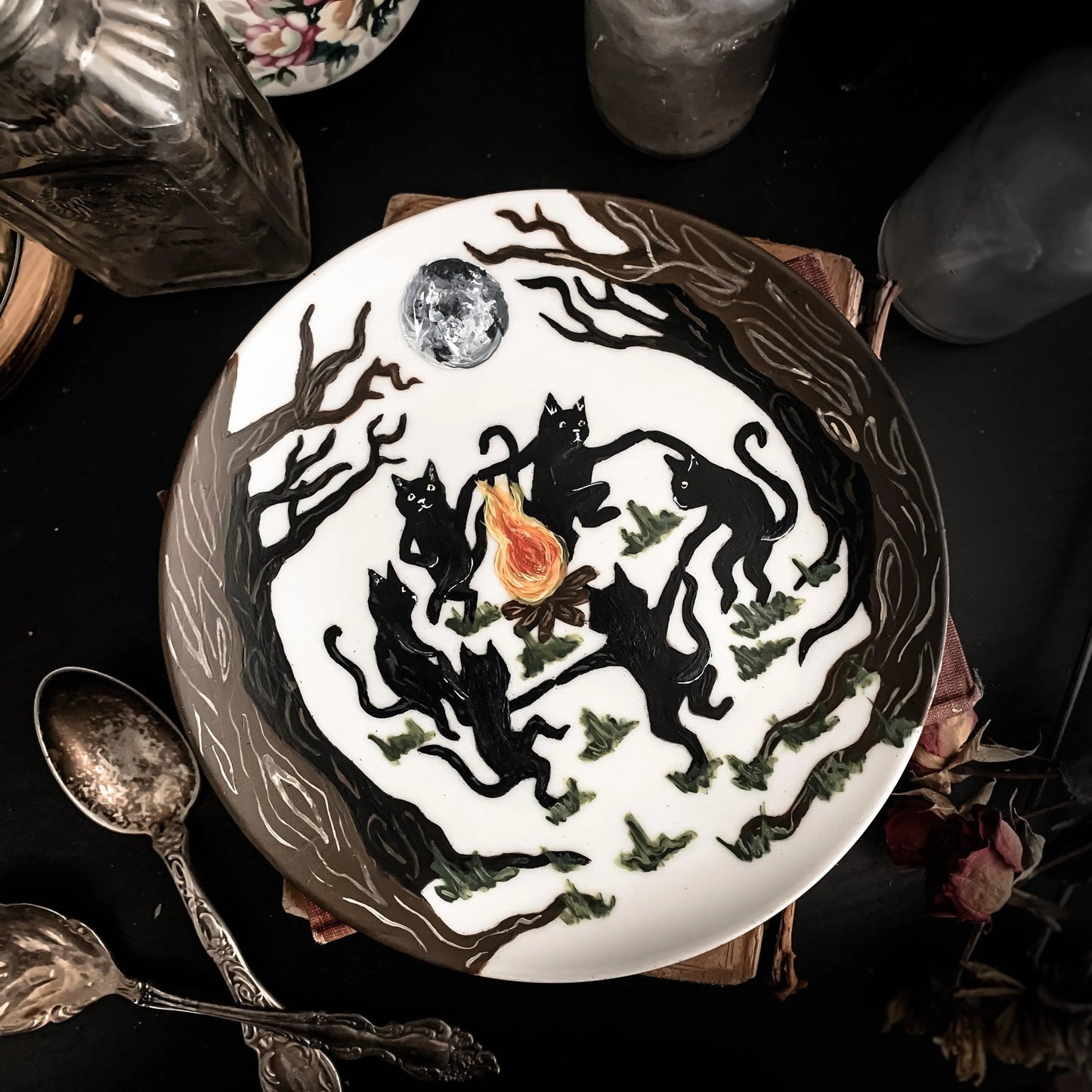 A decorative white plate featuring black cats dancing around a fire in the forest below a glowing full moon.