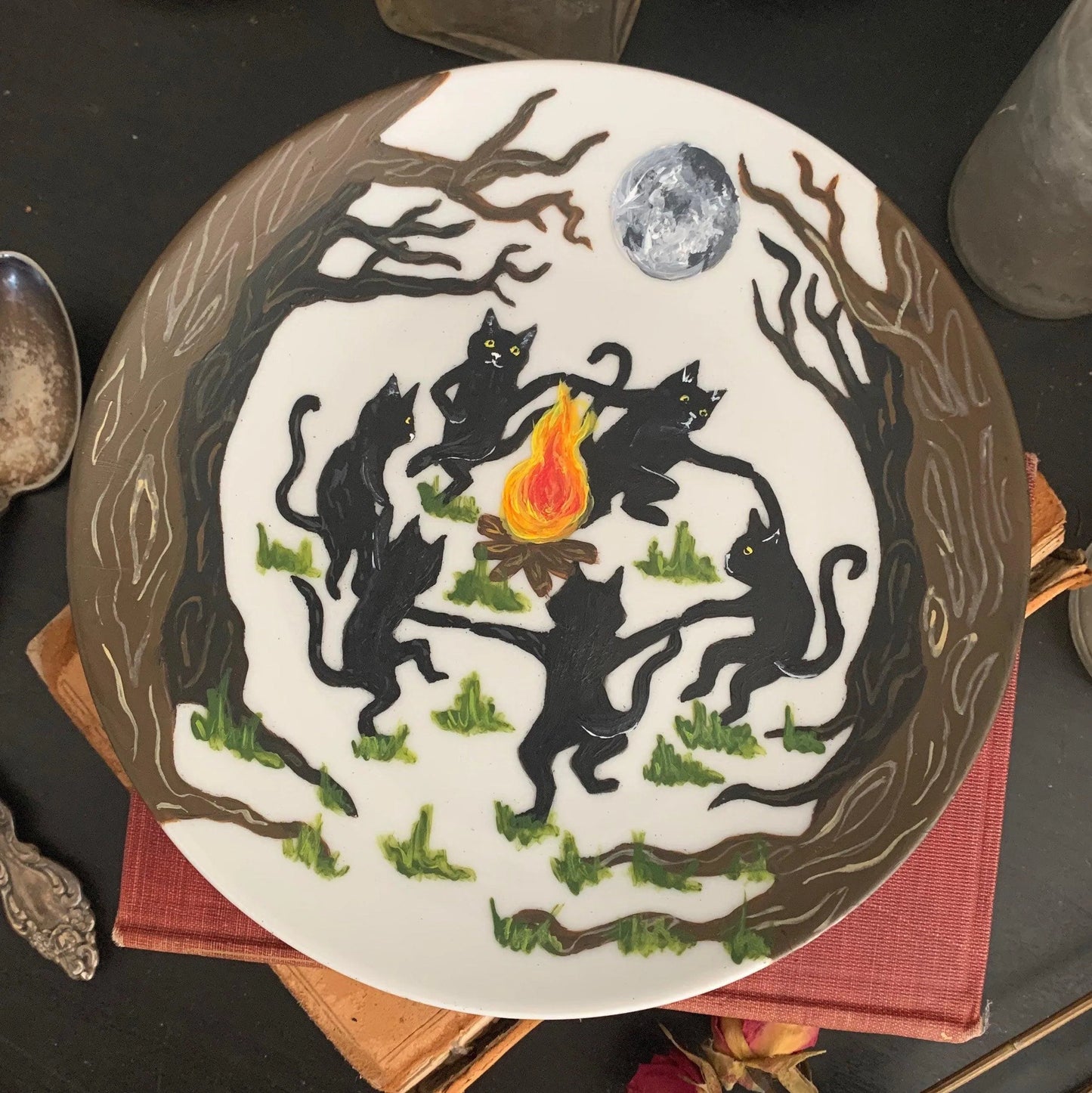 A decorative white plate featuring black cats dancing around a fire in the forest below a glowing full moon.