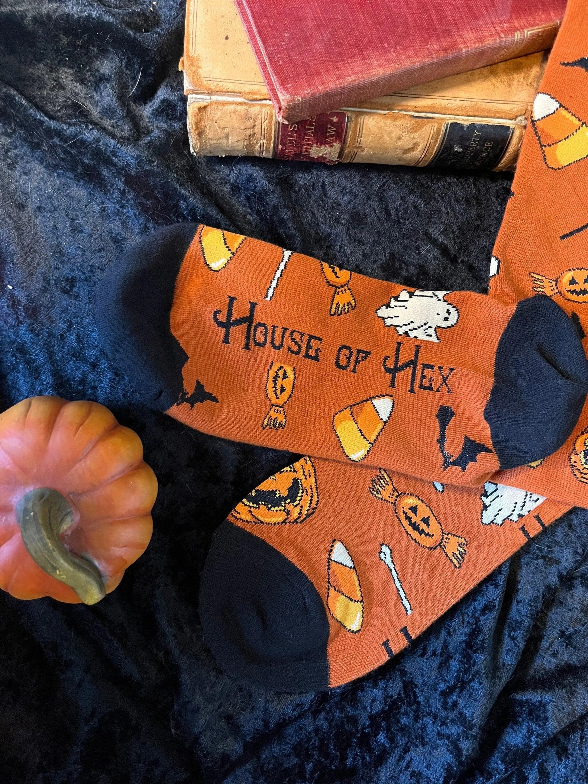 Halloween socks with blacks on the toes, heel, and ankle section featuring a pattern of pumpkins, candy corn, ghosts, bones, and bats. On the bottom is says "House of Hex".
