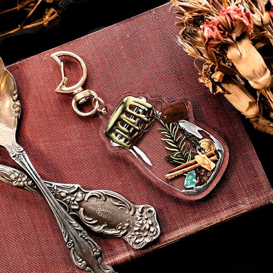 A decorative keychain displaying a small jar filled with cinnamon, bay leaves, coins, cloves, citrine, aventurine, and rosemary with a gold top. 