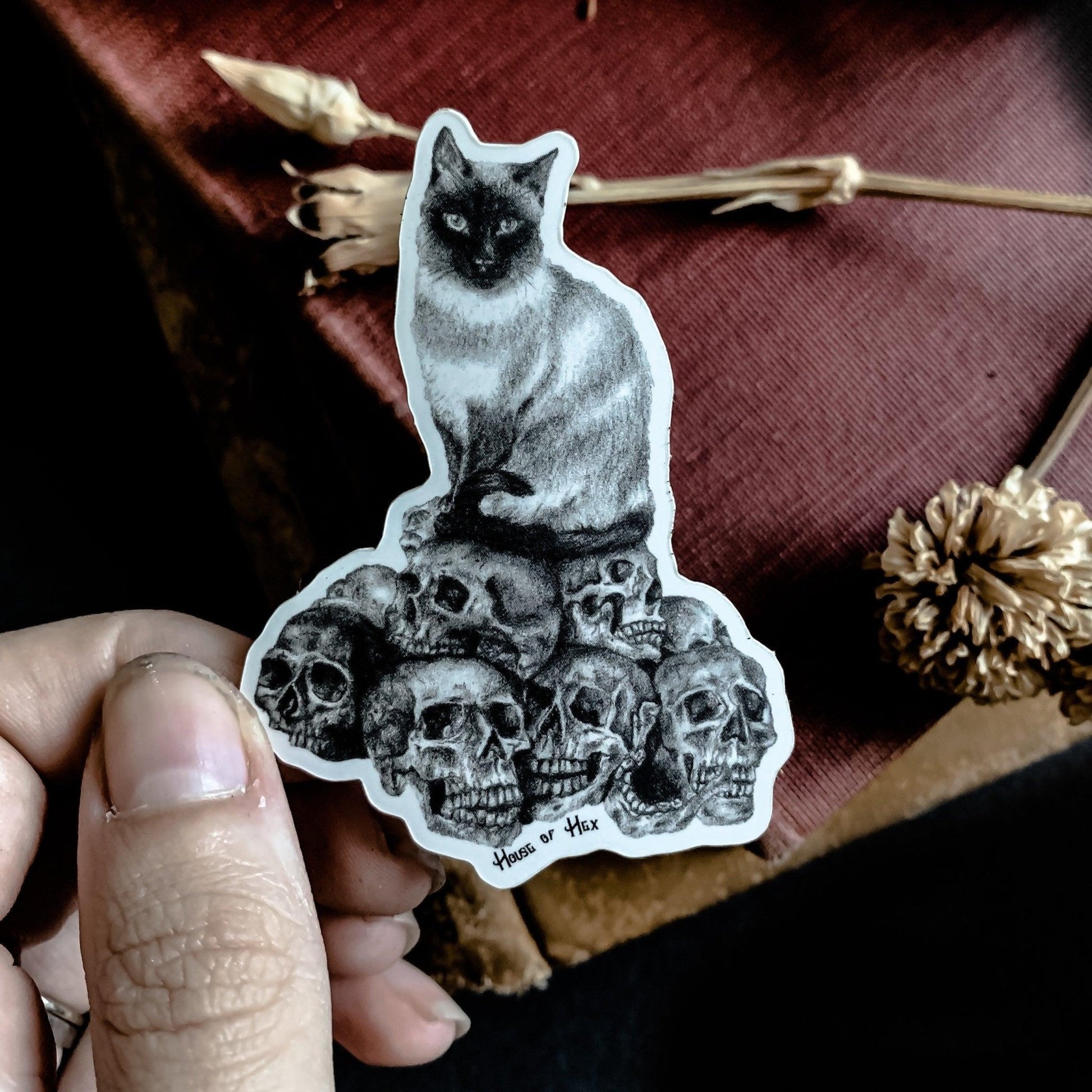 A black and white sticker with a drawing of a cat perched atop a collection of skulls. 