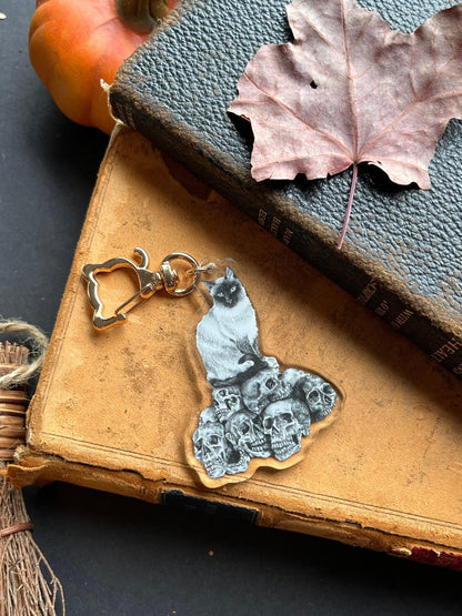 A black and white keychain with a drawing of a cat perched atop a collection of skulls with a golden cat-shaped clasp.